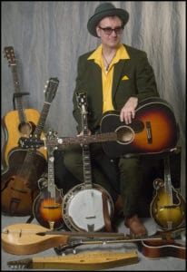 Photograph of Buddy in a hat and yellow shirt, with about 8 or more instruments.